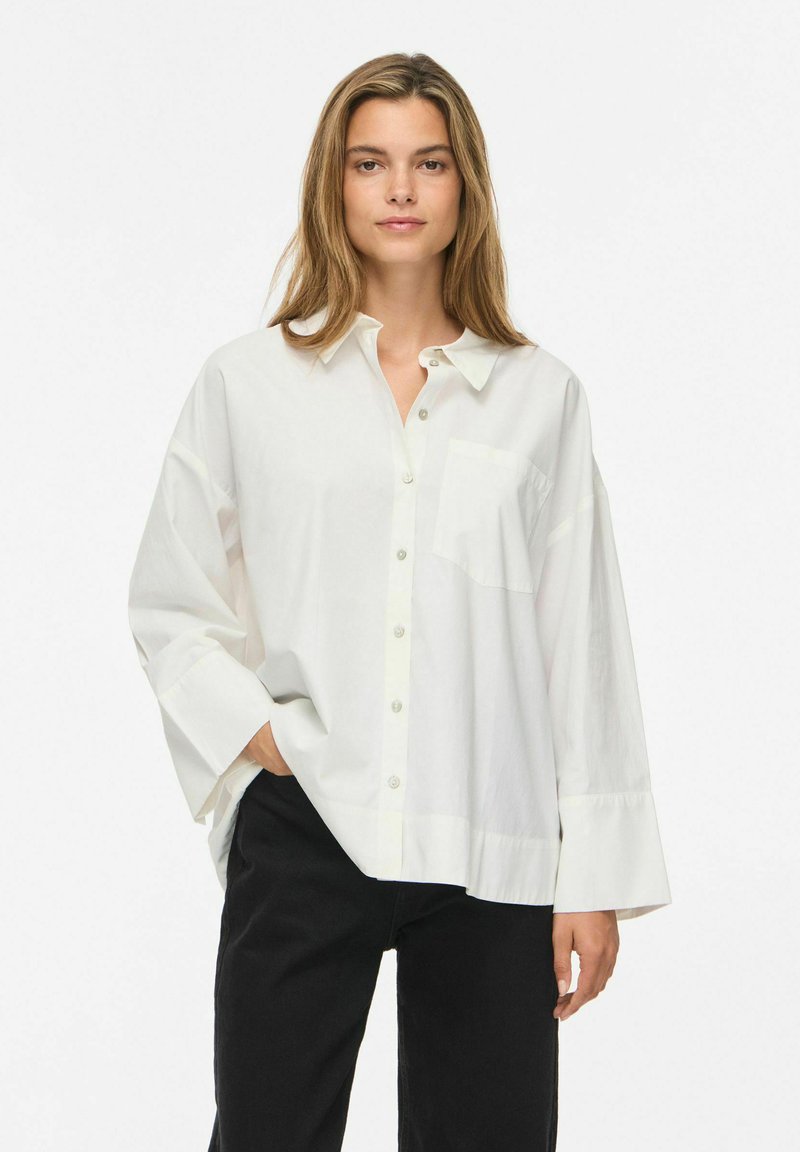 Femme aux longs cheveux portant une chemise blanche ample boutonnée et un pantalon noir, debout sur un fond blanc uni.