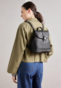 Svart läderryggsäck med klaffstängning, justerbara remmar och ett handtag på toppen. Slät yta, minimalistisk design och subtila hårdvaruaccents.