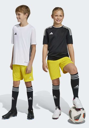 Two children wearing soccer gear: a white short-sleeve shirt, black shirt, yellow shorts, black socks with stripes, and cleats. Soccer ball present.