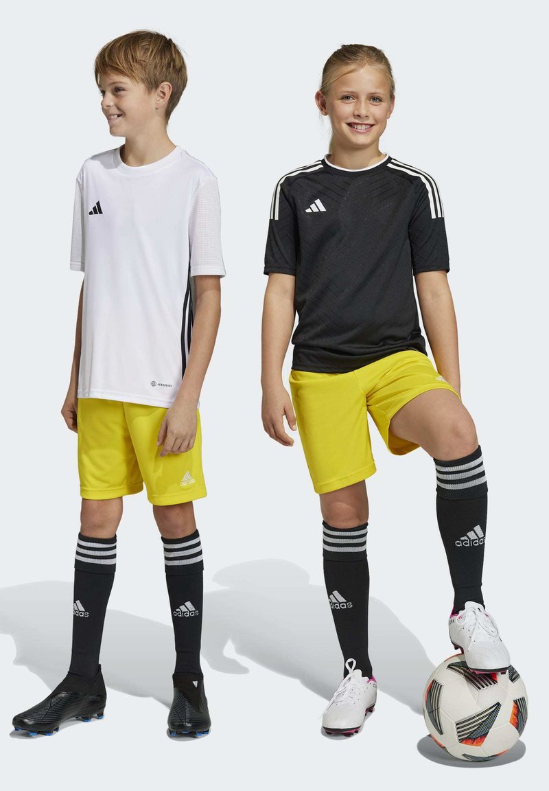 Twee kinderen in voetbalkleding: een wit T-shirt met korte mouwen, een zwart shirt, gele shorts, zwarte sokken met strepen en voetbalschoenen. Een voetbal aanwezig.