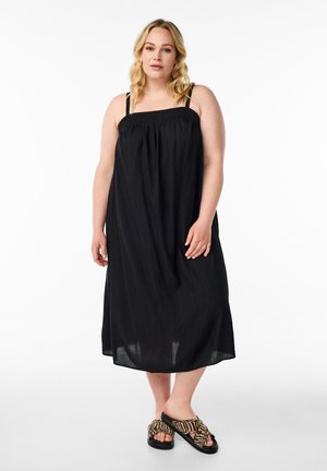 Schwarzes, ärmelloses Maxikleid mit gerafftem Oberteil und geradem Schnitt. Getragen mit gemusterten Sandalen, die eine offene Zehenpartie und ein überkreuztes Riemendesign aufweisen.