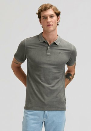 Giovane uomo con capelli ricci che indossa una maglietta polo grigia e jeans azzurri, in piedi con le mani dietro la schiena su uno sfondo semplice.