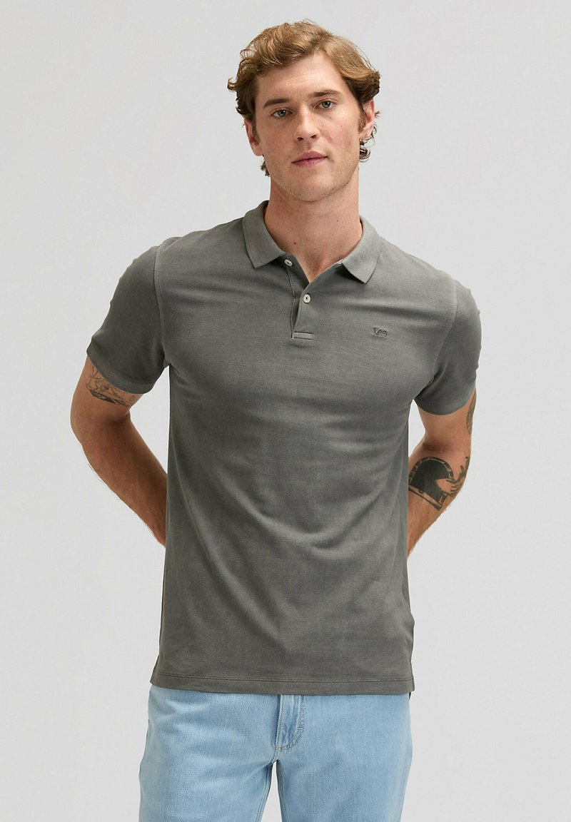 Giovane uomo con capelli ricci che indossa una maglietta polo grigia e jeans azzurri, in piedi con le mani dietro la schiena su uno sfondo semplice.