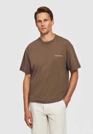 Mężczyzna w brązowej koszulce z krótkim rękawem z napisem "PAKO LORENTE", w połączeniu z jasnobeżowymi spodniami, stoi z jedną ręką w kieszeni.