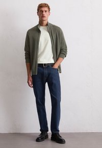 Lichtgewicht olijfgroene ritsjack over een wit shirt, gecombineerd met donkerblauwe jeans en zwarte veterschoenen. Eenvoudig ontwerp en aangesloten snit.
