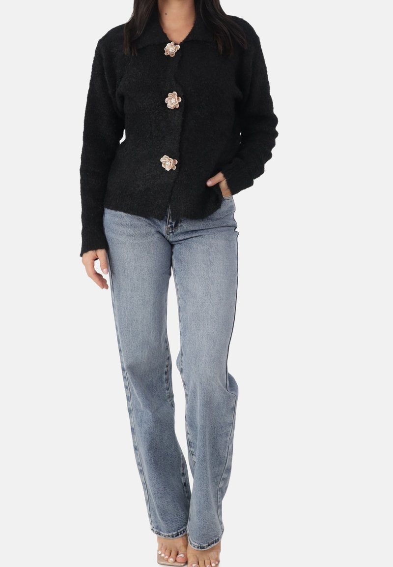 Femme portant un cardigan noir duveteux avec des boutons en forme de fleur et un jean bleu clair à jambes droites, debout avec une main dans la poche.