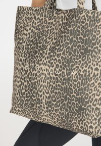 Sac fourre-tout au motif léopard en tissu, avec un fond beige et des taches noires, et le texte "ATHLECIA" en blanc. Design structuré avec deux poignées.