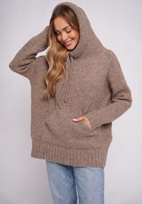 Brązowa dzianinowa bluza z kapturem z wysokim, prążkowanym kołnierzem, detalem ze sznurkiem i kieszenią z przodu. Luźny krój i miękka faktura, w połączeniu z jasnymi jeansami.