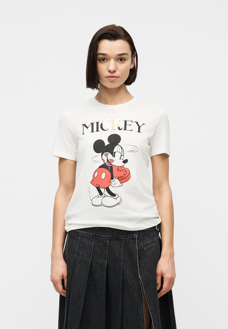Jeune femme aux cheveux courts et foncés portant un t-shirt blanc à motif Mickey Mouse et une jupe plissée noire taille haute avec une fente à l'avant.