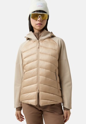 Veste isolée beige avec un devant matelassé, des manches lisses, une capuche, une fermeture éclair frontale et des poches latérales. Portée avec un bonnet en tricot beige et de grandes lunettes de soleil.