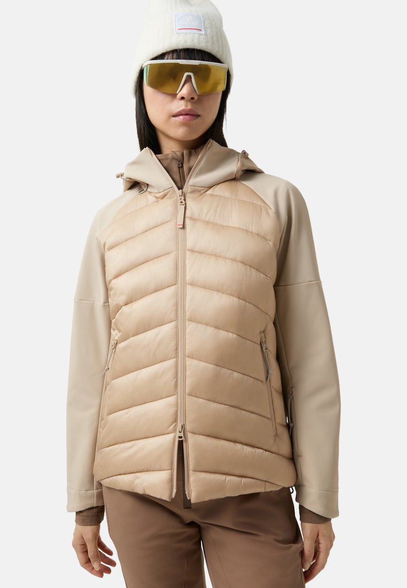 Beżowa kurtka ocieplana z pikowanym przodem, gładkimi rękawami, kapturem, zamkiem błyskawicznym z przodu oraz bocznymi kieszeniami. Noszona z beżową dzianą czapką i dużymi okularami przeciwsłonecznymi.