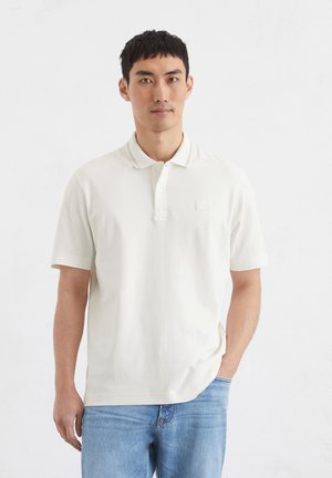 Light beige polo shirt with short sleeves, classic collar, and a subtle logo on the chest, paired with light blue denim jeans.