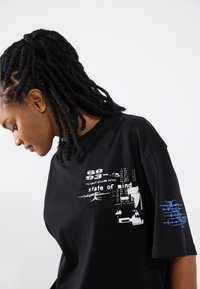 Black graphic t-shirt with large white and blue prints featuring text and abstract designs on the front and right sleeve. Short sleeves, relaxed fit.