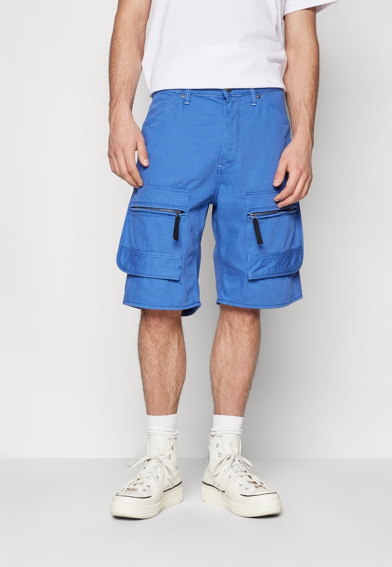 Homme portant un short cargo bleu avec poches à fermeture éclair et rabats, chaussettes blanches, baskets montantes blanches et un t-shirt blanc, debout devant un fond uni.
