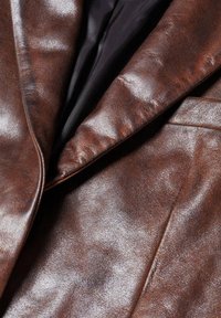 Ruskea nahkatakki kiiltävällä pinnalla, jossa on neulonnokka ja musta sisävuori. Pintarakenteen yksityiskohdat näkyvät selvästi.