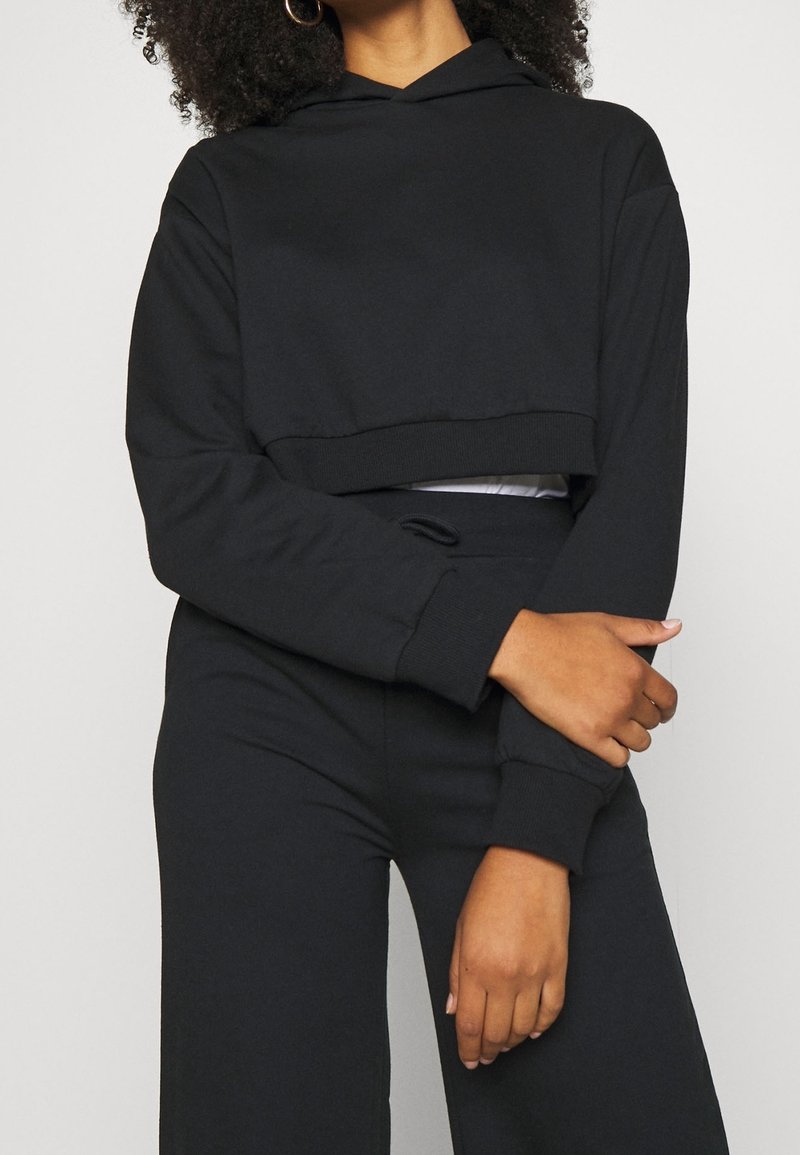 Femme portant un sweat à capuche court noir et un pantalon noir assorti, bras croisés à la taille sur un fond clair uni.