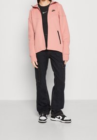 Rosa Kapuzenpullover mit Reißverschluss und hohem Kragen, kombiniert mit einem schwarzen, eng anliegenden Shirt, dunklen Cargo-Hosen und schwarz-weißen Sneakern.