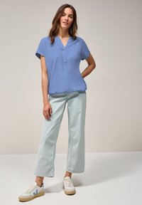 Blouse bleu clair à manches courtes avec un col en V et une texture douce, associée à un pantalon en denim clair à jambes larges et des baskets beiges avec des accents bleus.