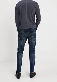 Jeans skinny azul oscuro con un ligero desgaste, cinco bolsillos y costuras dobles. Combinados con una camisa gris de manga larga y zapatos negros.