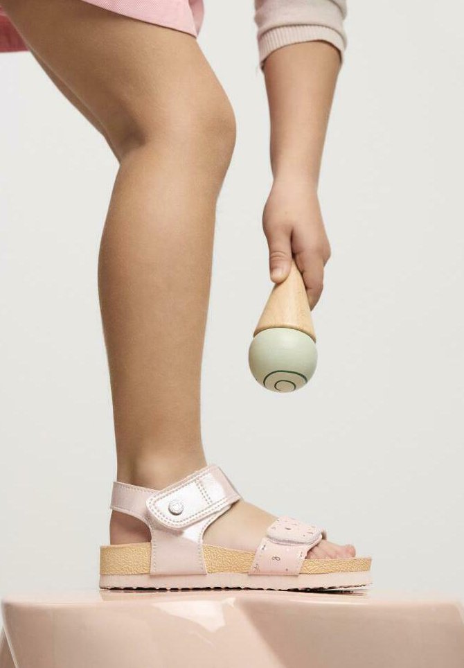 Sandalias de color rosa claro con una tira brillante y una suela de corcho texturizada. Presenta una tira decorativa con pequeños recortes. Mano de un niño sosteniendo un juguete.