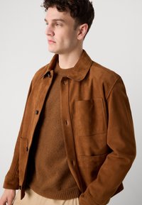 Brown suede jacket with a classic collar, front button closure, and a patch pocket. Worn over a brown knit sweater.