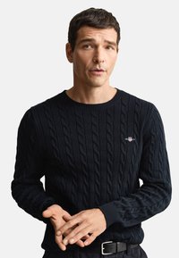 Marineblauwe kabelgebreide trui met een ronde hals en geribbelde manchetten, met een klein geborduurd logo op de borst. De structuur lijkt zacht en dik.