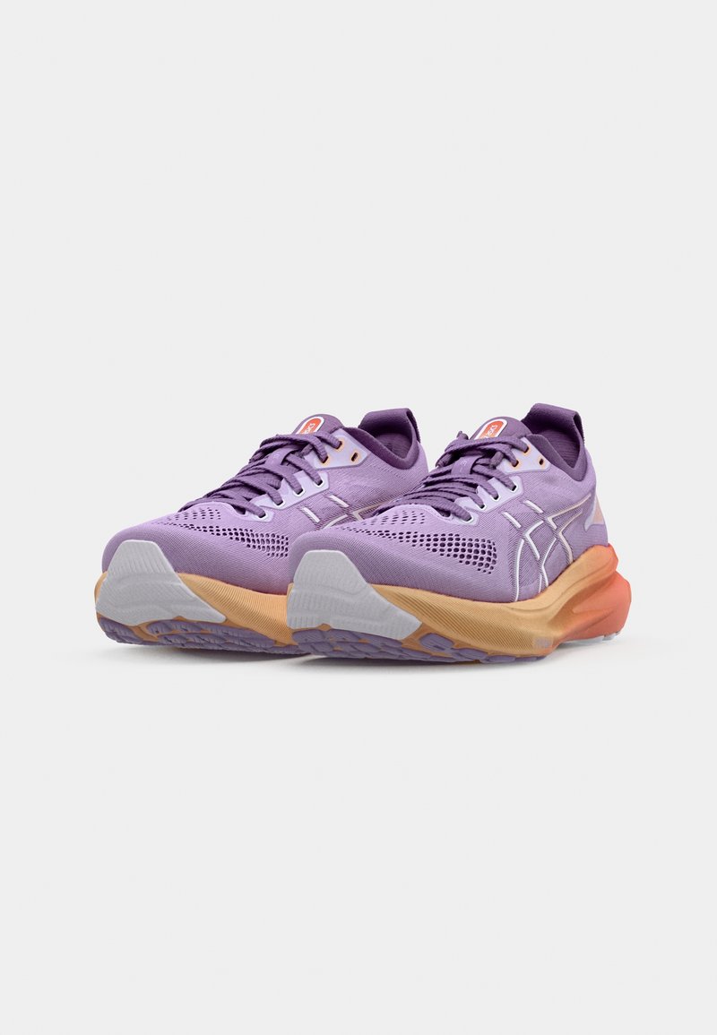 Zapatillas deportivas moradas con parte superior de malla texturizada, suela exterior en naranja y gris contrastante, y acentos en blanco. Presentan cordones y logo.