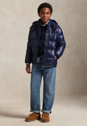 Junge steht und trägt eine marineblaue Steppjacke, ein gestreiftes Hemd, einen dunklen Pullover, blaue Jeans und braune Schnürschuhe vor neutralem Hintergrund.