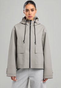 Veste imperméable gris clair avec capuche, fermeture éclair à l'avant, deux grandes poches et manches larges. Matériau texturé avec cordons de serrage ajustables.