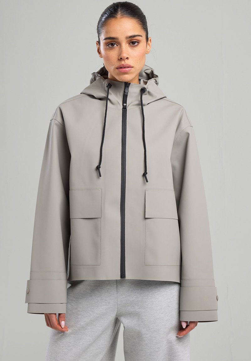 Veste imperméable gris clair avec capuche, fermeture éclair à l'avant, deux grandes poches et manches larges. Matériau texturé avec cordons de serrage ajustables.