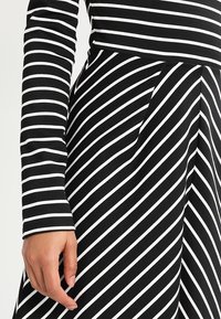 Robe à rayures noires et blanches avec des manches longues et un corsage ajusté, présentant un motif diagonal sur la jupe et un tissu doux et texturé.