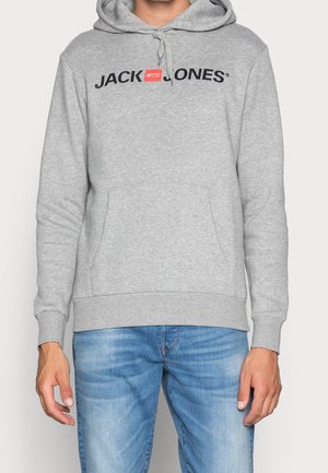 Man som bär en grå hoodie med "JACK & JONES"-logotyp i svart och rött på bröstet, i kombination med blå jeans, stående mot en enfärgad bakgrund.
