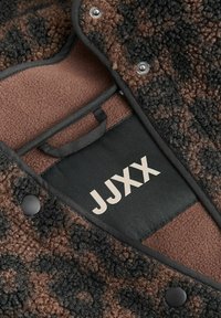 Brun og svart fleecejakke med teksturert utside, glatt innerfôr, metalliske trykknapper og en etikett med teksten "JJXX".