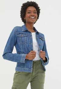 Smiling woman wearing a blue denim jacket, white shirt, and olive green pants, standing against a plain light background.