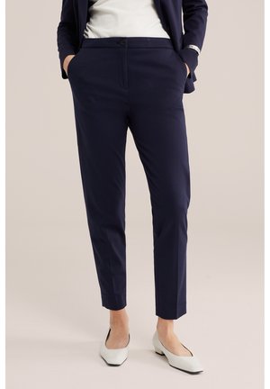 Personne portant un pantalon bleu marine sur mesure, une blouse blanche, un blazer bleu marine et des chaussures blanches à petits talons, debout les mains dans les poches.