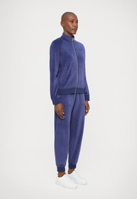 Osoba stoji u usklađenom plavom velour zip-up dukserici i trenirkama s bijelim tenisicama, naslonjena na svijetlu jednostavnu pozadinu.