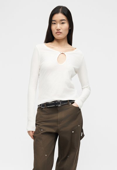 Νεαρή γυναίκα με μακριά σκούρα μαλλιά που φοράει λευκό μακρυμάνικο τοπ με άνοιγμα στο μπροστινό μέρος και καφέ παντελόνι με μαύρη ζώνη με τρουκς.