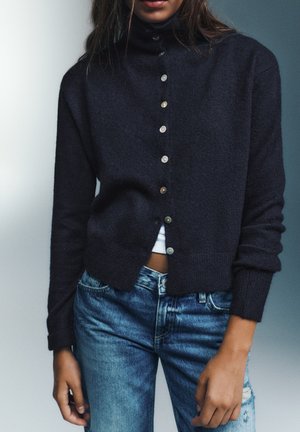 Personne portant un cardigan bleu marine foncé boutonné sur un haut blanc avec un jean bleu, debout devant un fond neutre.