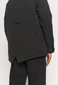 Doudoune noire en tissu texturé, avec un col haut, un ourlet aéré et des accents de boutons-pression aux poignets, présentant une coupe décontractée.