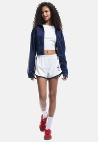 Mørkeblå zip-up jakke, hvid crop top og hvide træningsshorts med mørke kanter. Røde sneakers med hvide detaljer og tykke hvide sokker.