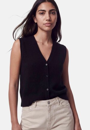 Femme aux longs cheveux foncés portant un gilet noir sans manches boutonné et un pantalon beige clair sur un fond blanc uni.