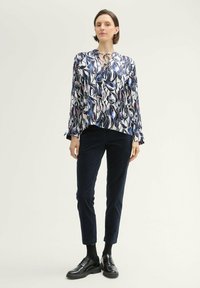 Blouse à manches longues avec un imprimé abstrait multicolore et un col à lien en forme de clé, associé à un pantalon slim noir et des chaussures à lacets noires.