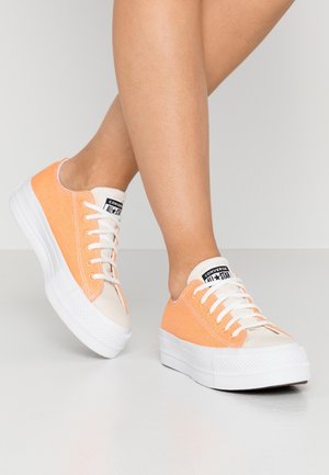 Personne portant des baskets Converse All Star basses orange et beige avec des lacets blancs et des semelles blanches épaisses sur un fond neutre.