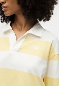 Γυναίκα που φοράει κίτρινη και λευκή ριγέ ράγκμπι μπλούζα με ανοιχτό γιακά και λογότυπο New Balance στο στήθος, με εμφανή σγουρά μαλλιά.