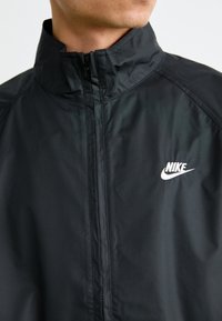 Nærbillede af en person iført en sort Nike-jakke med en lynlåset krave og et hvidt Nike-logo på brystet.