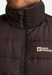Gevoerde zwarte jas met hoge kraag, volledige rits en Jack Wolfskin-logo. Glad textuur en stevige stof met gestikte panelen.