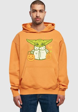 A narancssárga pulóver nagy grafikával rendelkezik egy zöld bőrű karakterről, aki két halloween-i tököt tart a kezében. Elöl zsebe és kapucnija van.