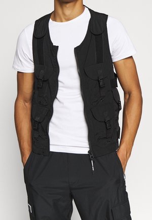 Mann trägt eine schwarze Utility-Weste mit mehreren Taschen mit Schnallen über einem weißen T-Shirt und schwarzen Cargo-Hosen, Hände in den Taschen.