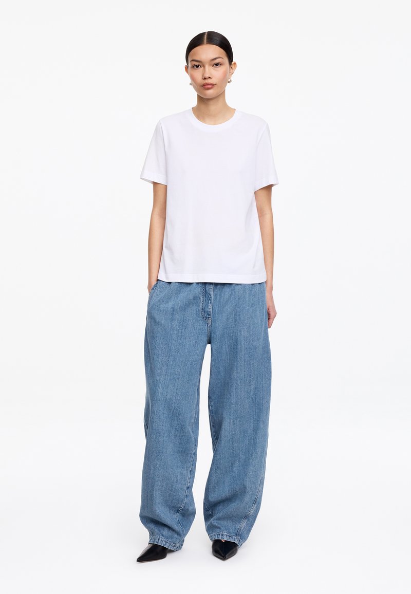 Femme debout de face, portant un t-shirt blanc uni à manches courtes, un jean en denim bleu ample et des chaussures noires pointues sur fond blanc.