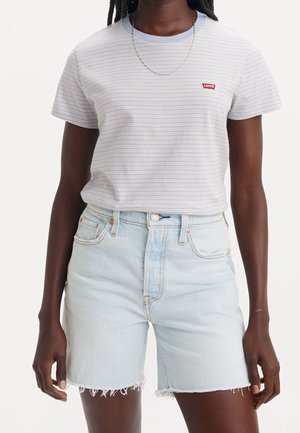 Osoba ubrana w jasnoniebieski pasiasty t-shirt Levi’s i jasne szorty jeansowe z postrzępionymi brzegami na jednolitym białym tle.